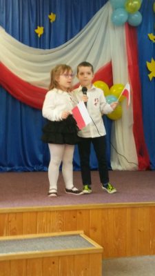 Dziewczynka z chłopcem stoją na scenie, kołyszą się. W rękach trzymają polskie flagi.
