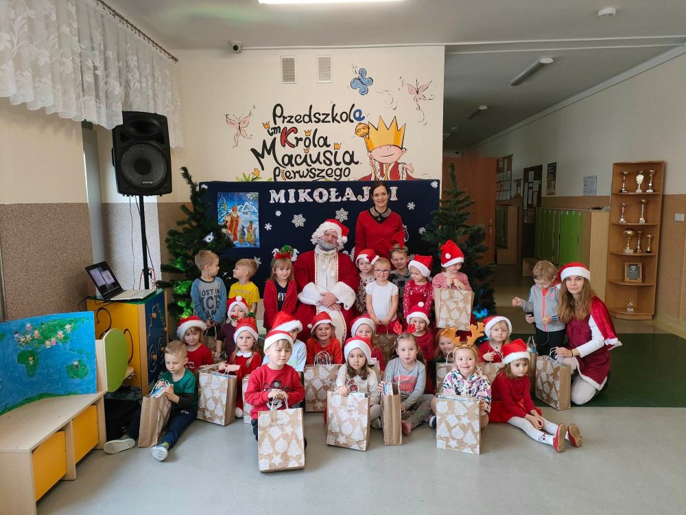 Dzieci z grupy Śmieszki pozują do zdjęcia ze Świętym Mikołajem. Przedszkolaki trzymają swoje prezenty, a w tle widać mikołajową dekorację.