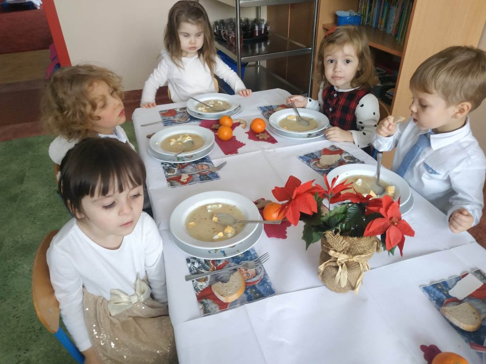 Przy wigilijnym stole siedzi 5 dzieci. Są elegancko ubrane. Za stole leżą talerze z zupą i dekoracja.