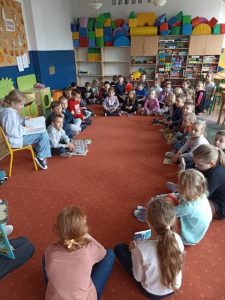 Dzieci siedzą w kręgu na dywanie i słuchają uczennicy czytającej książkę.