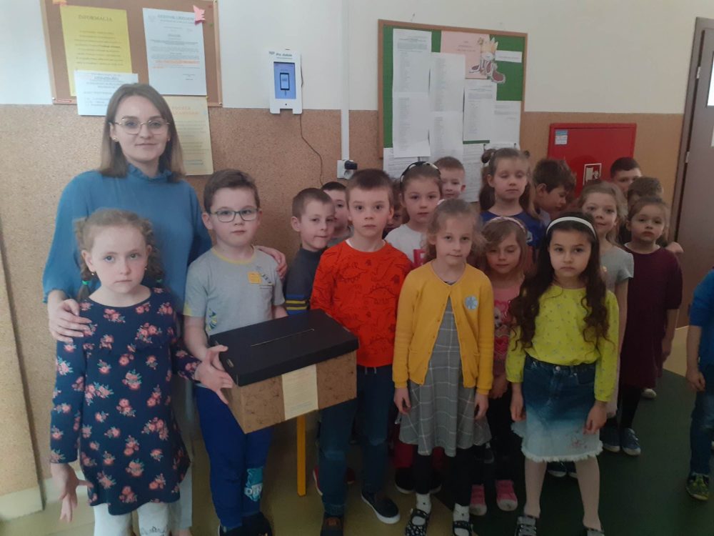 Nauczycielka wraz z 18 dzieci. Dziecko trzyma pudełko z kartkami wielkanocnymi.