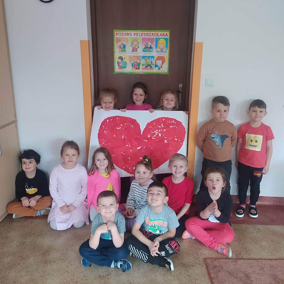 13 dzieci pozuje na tle drzwi. 3 z nich trzyma wielki plakat z czerwonym sercem. W tle biała ściana i szafa.