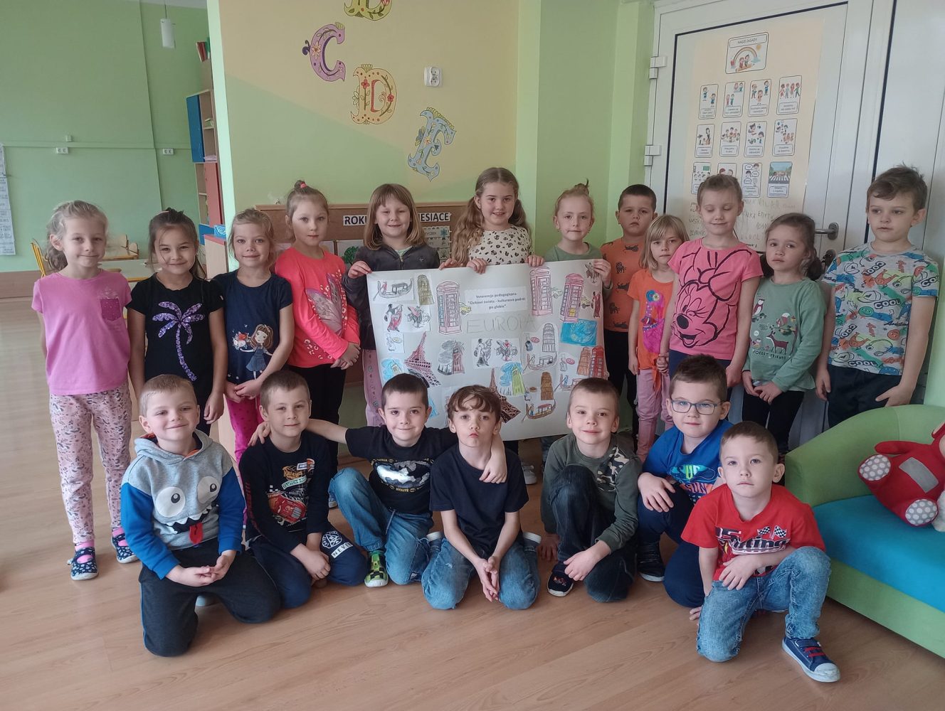 Na zdjęciu widać 19 dzieci na tle kolorowej ściany. Dzieci prezentują plakat przedstawiający informacje na temat Europy. W tle widać drzwi, ścianę i tablicę.