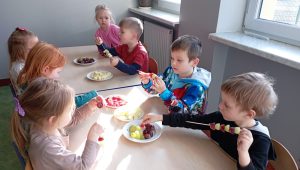 Dzieci siedzą przy stole i robią szaszłyki z owoców i warzyw.