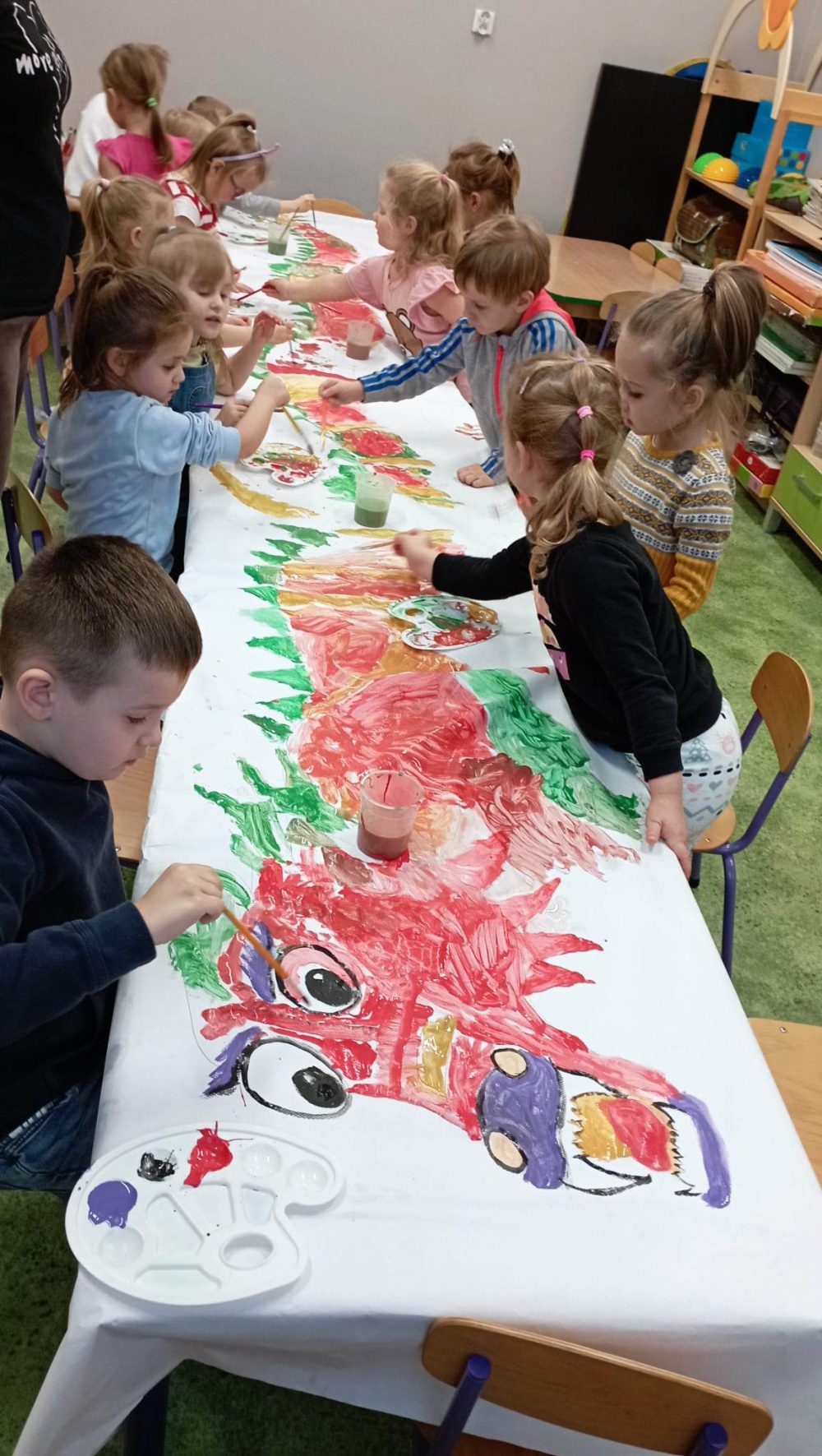 Dzieci siedzą przy stole i malują długiego smoka farbami.