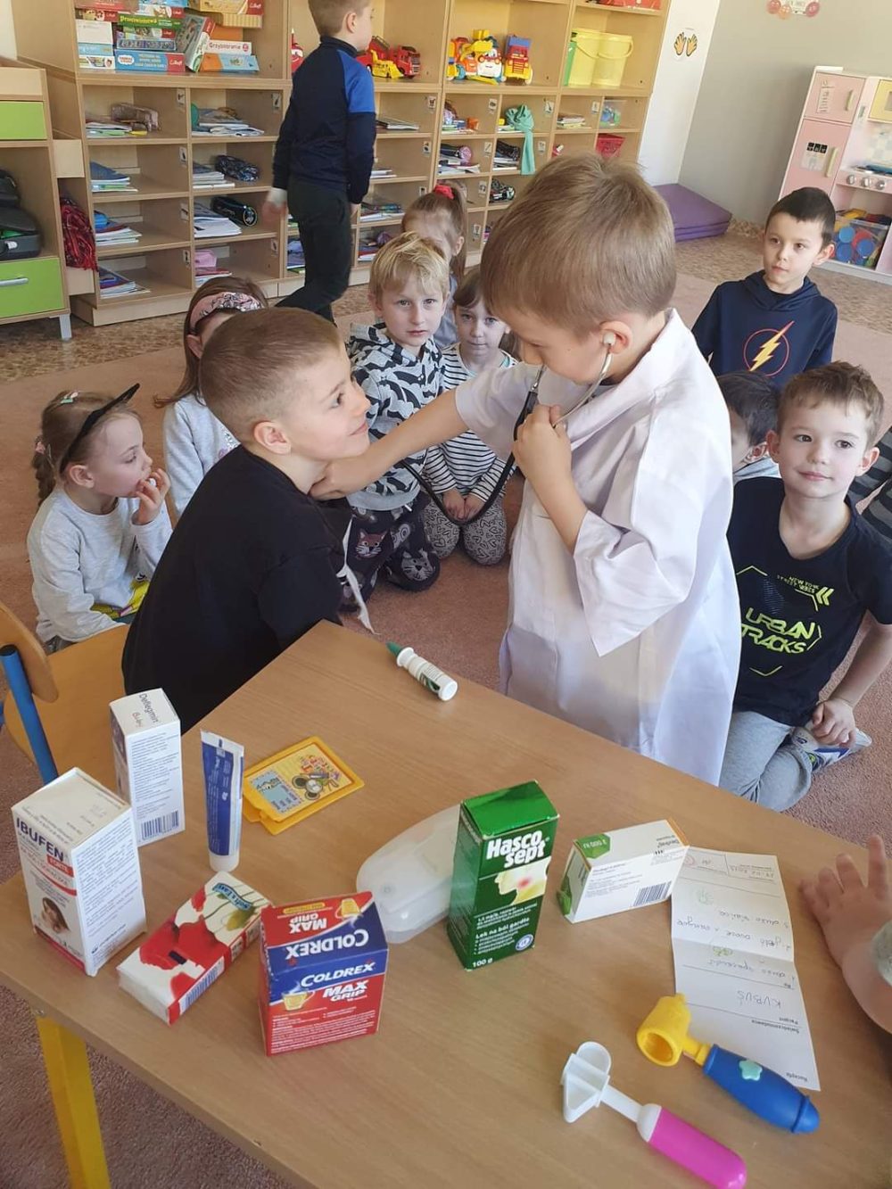 chłopiec przebrany w biały fartuch lekarski bada stetoskopem swojego kolegę z grupy, który przybrał rolę pacjenta.