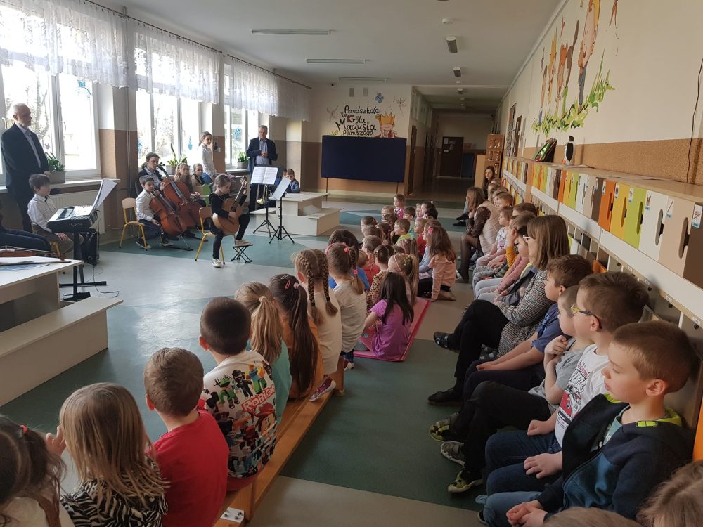 Grupa przedszkolaków ogląda występ dziewczynki grającej na gitarze. Za dziewczynką siedzą nauczyciele i dzieci ze szkoły muzycznej. Trzymają w dłoniach instrumenty.