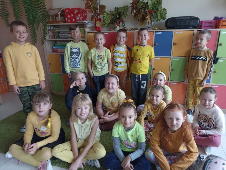 Na zdjęciu widać 15 osobową grupę dzieci ubranych w stroje w żółtych barwach. Sześciu chłopców stoi, a pozostali siedzą przed nimi na dywanie. Wszyscy uśmiechają się i pozują do zdjęcia. W tle widać kolorowe szafki i jesienne dekoracje z liści.