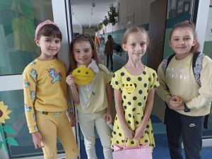 Na zdjęciu widać cztery stojące obok siebie dziewczynki. Wszystkie uśmiechają się. Dwie z nich mają na żółtych bluzkach emotikony przedstawiające uśmiech. W oddali widać szkolny korytarz.