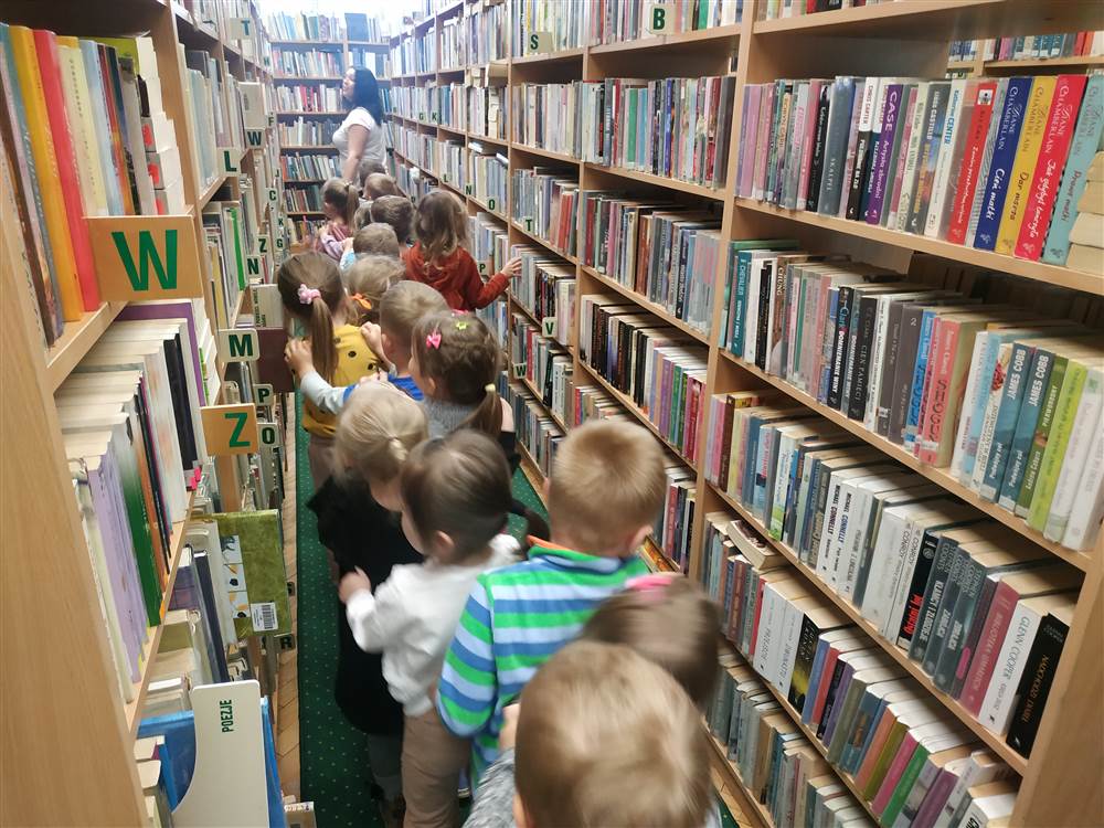 Grupa dzieci wraz z dorosłym zwrócona tyłem do aparatu stoi pomiędzy regałami wypełnionymi książkami.