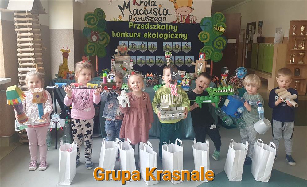 Dzieci z grupy Krasnale stojące ze swoimi pracami konkursowymi przy tablicy z napisem Przedszkolny Konkurs Ekologiczny: Zrób coś z niczego.