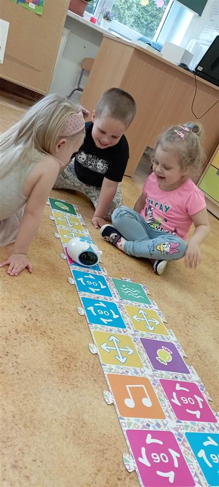 Trójka dzieci siedzi na podłodze i programuje robota. Między nimi leży plansza do programowania z puzzli i robot.
