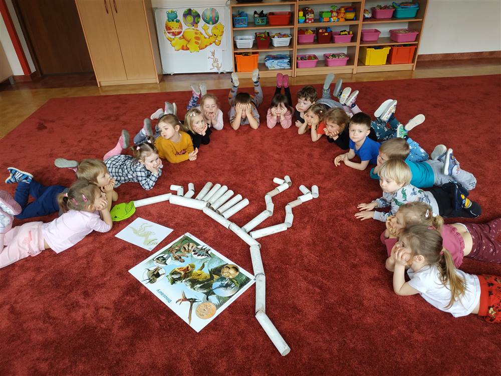 Grupa dzieci leży w półkolu na dywanie w leżeniu przodem. Na dywanie znajduje się szkielet dinozaura ułożony z tekturowych rolek oraz plansza z dinozaurami.