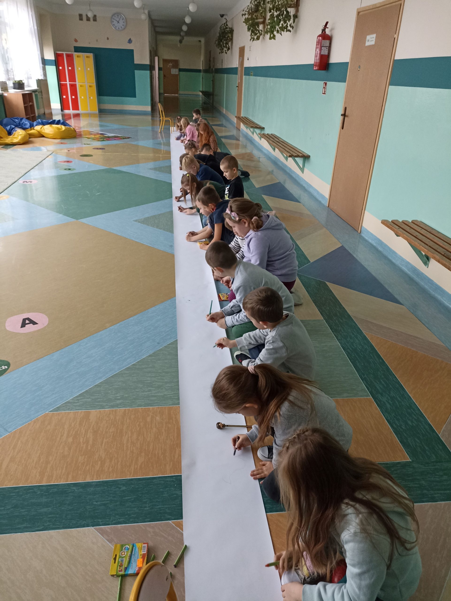 Na korytarzu szkolnym grupa dzieci siedzi w rzędzie i rysuje na długiej kartce papieru.