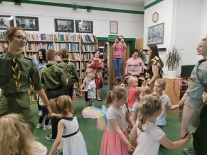 Dzieci, harcerze i panie tańczą razem w kołach. Pani bibliotekarka prowadzi zabawę. W tle widać półkę z książkami. Pani fotograf robi zdjęcie.