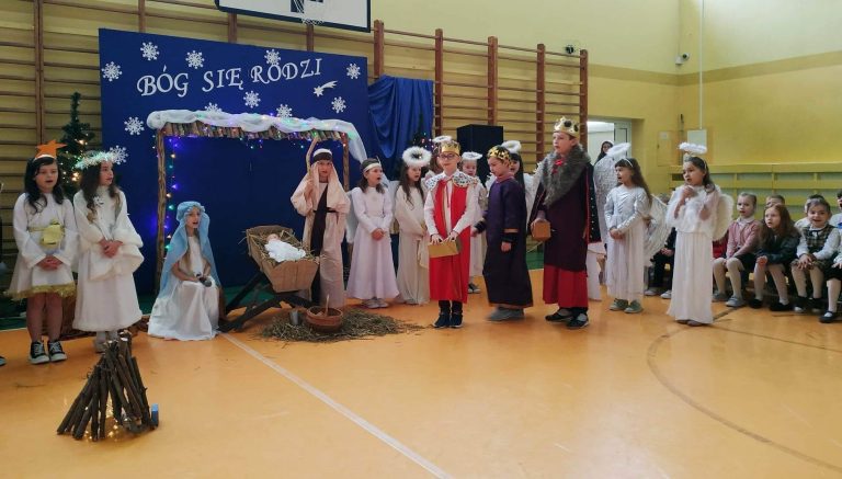 Na tle niebieskiej dekoracji dziewczynkasiedzi, a chłopiecstoi przy żłóbku.Po obu stronach dzieci przebrane w stroje aniołów i królów.