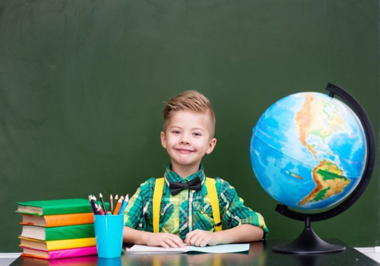 Chłopiec siedzi przy stoliku. Na stole po jego prawej stronie leżą książki i stoi kubek z kredkami, a po lewej stoi globus. W tle widać tablicę szkolną.