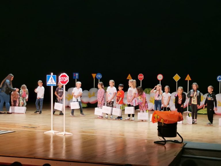 Grupa dzieci stoi w rzędzie na scenie. Dzieci trzymają torby prezentowe. W tle widać różne modele znaków drogowych.