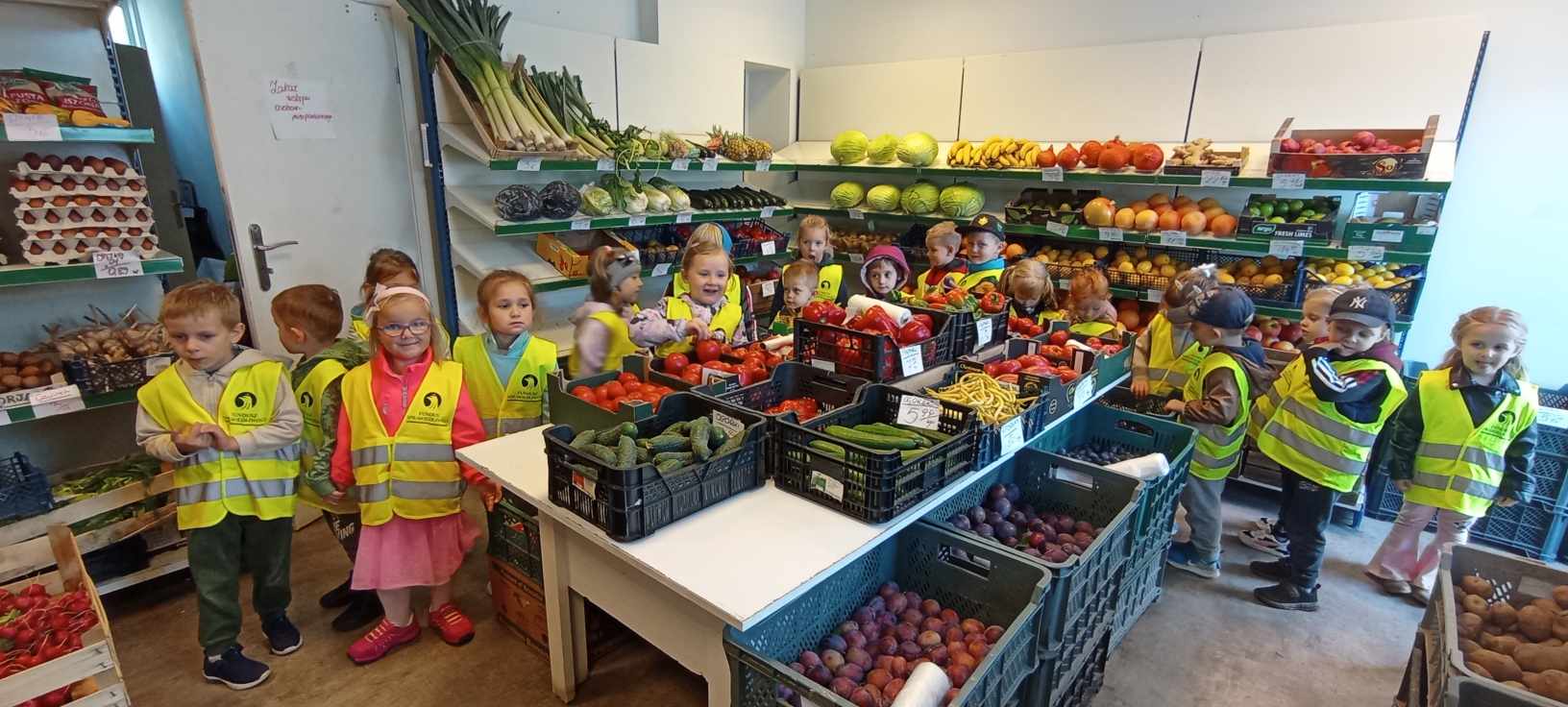 Grupa dzieci stoi w sklepie między regałem a stoiskiem z warzywami. Do koła leży dużo warzyw i owoców w skrzynkach.