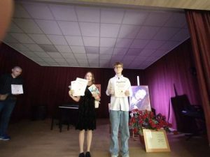Na zdjęciu widzimy dziewczynkę i chłopca w strojach galowych, trzymających dyplomy i nagrody. Uczniowie stoją na scenie. W tle portret pisarki Bronisławy Betlej i duży bukiet czerwonych róż.