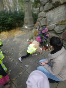 Dziecko kładzie znicz pod pomnikiem na cmentarzu. Nauczyciel asystuje dziecku dla bezpieczeństwa. Pozostałe dzieci obserwują pomnik.