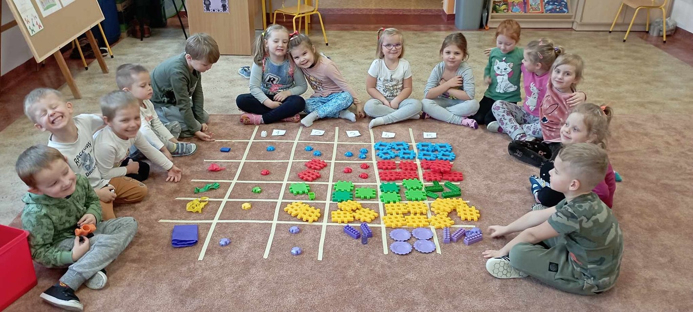 Grupa dzieci siedzi na dywanie wokół ułożonego kodowania - kratownicy z cyframi, kolorami i klockami w odpowiedniej liczbie.
