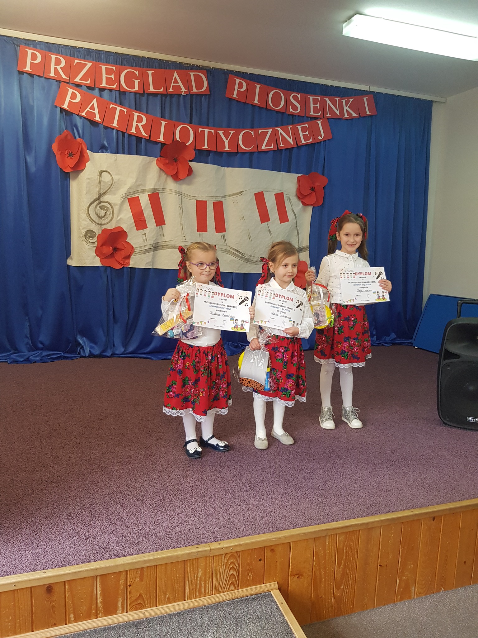 Trzy dziewczynki stoją na scenie w strojach góralskich i pozują do zdjęcia trzymając w ręce dyplomy. W tle widać dekorację z napisem "Przegląd Piosenki Patriotycznej".