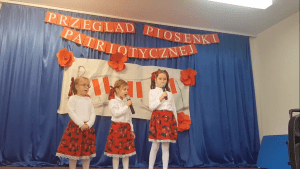 Trzy dziewczynki stoją na scenie w strojach góralskich i śpiewają piosenkę. W tle widać dekorację z napisem "Przegląd Piosenki Patriotycznej".