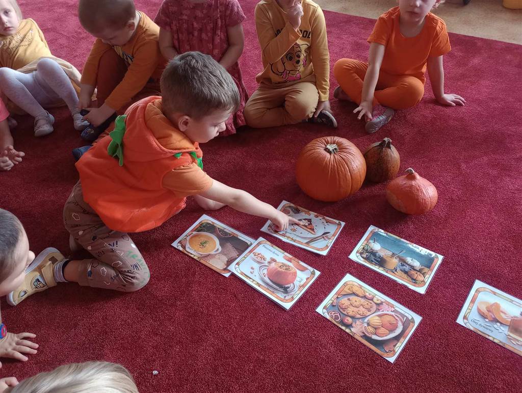 Grupa dzieci siedzi na dywanie. Chłopiec wskazuje na obrazek przedstawiający dynię.