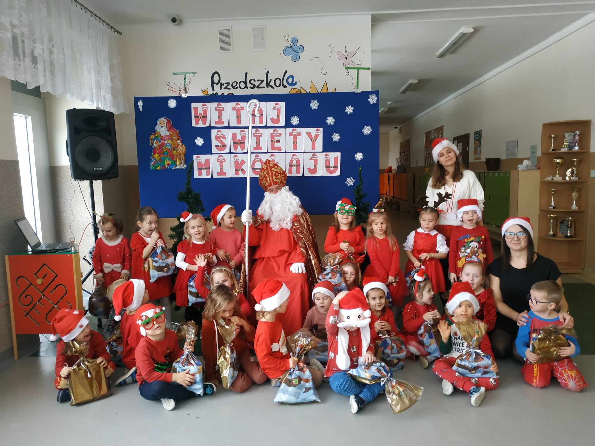 Grupa Śmieszki pozuje do zdjęcia z Mikołajem i nauczycielem. Dzieci w rękach trzymają prezenty. W tle widać mikołajkową dekorację.