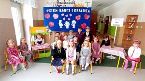 Grupa dzieci pozuje do zdjęcia. 11 dziewczynek siedzi na krzesłach, 6 chłopców stoi z tyłu, a jedna dziewczynka stoi w sklepiku. Dzieci i dekoracja jest w stylu PRL-u.