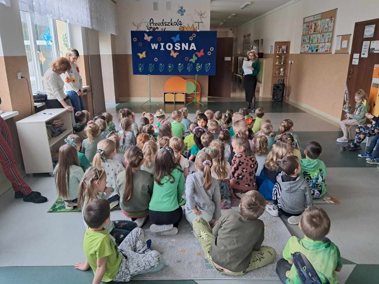 Przedszkolaki siedzą na dywanikach i słuchają przemówienia nauczyciela. Z tyłu widać wiosenną dekorację.