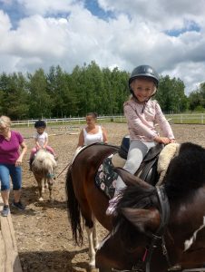 Na pierwszym planie dziewczynka jedzie na koniu, na drugim planie inne dziecko jedzie na koniu którego prowadzą dwie panie.