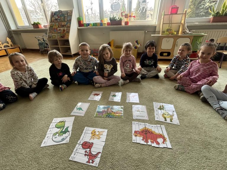 Grupa dzieci siedzi na dywanie w półkolu, a po środku leż ułozone puzzle.