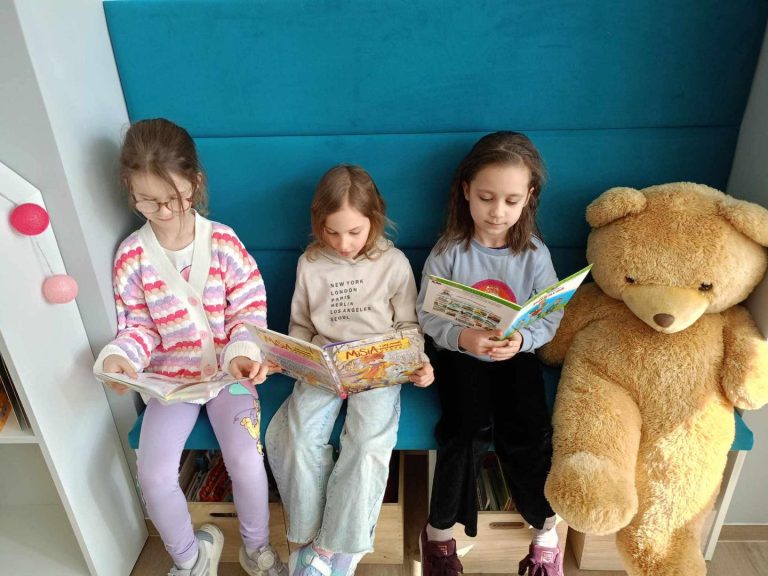 Trzy dziewczynki czytają książki siedząc na ławeczce. Obok leży duży miś.