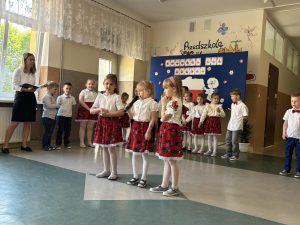 Dziewczynki mówią wiersze podczas apelu. W tle widać inne dzieci i dekorację.