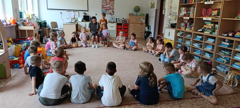 Grupa dzieci siedzi na dywanie i słucha bajki czytanej przez jedną z mam.