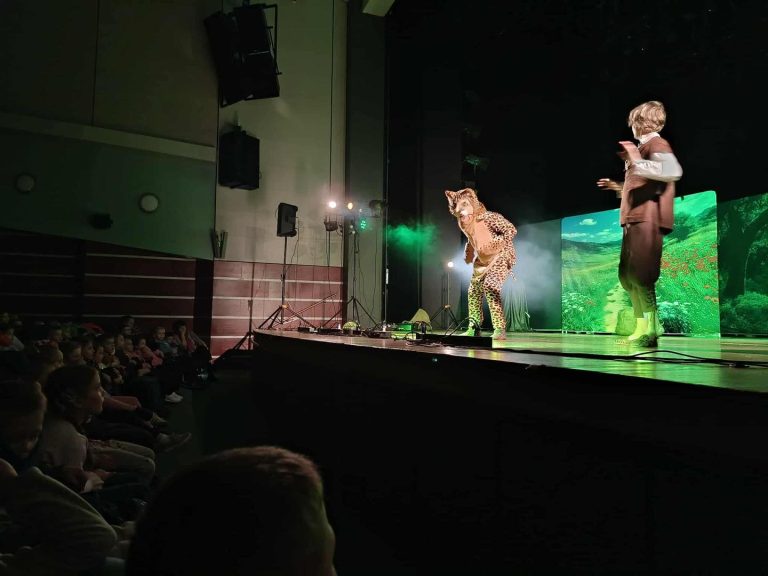 Na scenie widać aktorów w kostiumach odkrywających przedstawienie i dekorację. Z przodu widać widownię.