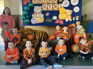 Grupa dzieci w raz z nauczycielem pozuje do zdjęcia. W tle widać dekorację na dzień dyni.