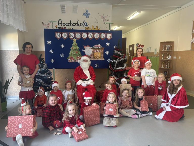 Grupa dzieci pozuje do zdjęcia z Mikołajem oraz z nauczycielami. W tle widać dekorację.