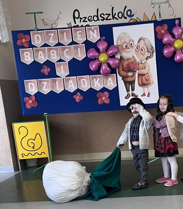 Dwoje dzieci przebranych za dziadków uczestniczy w przedstawieniu. Za nimi widać dekorację.