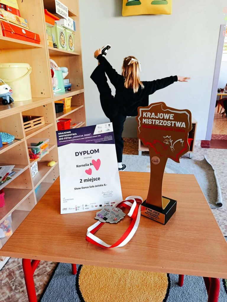 Na pierwsyzm planie widać dyplom, medal i statuetkę. W tle dziewczynka wykonuje figurę taneczną.